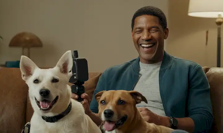 Un alegre actor, Denzel, rodeado de un perro juguetón, sentado en un sofá, riéndose con un amigo o una co-estrella, sosteniendo una cámara o micrófono, con una película en el fondo